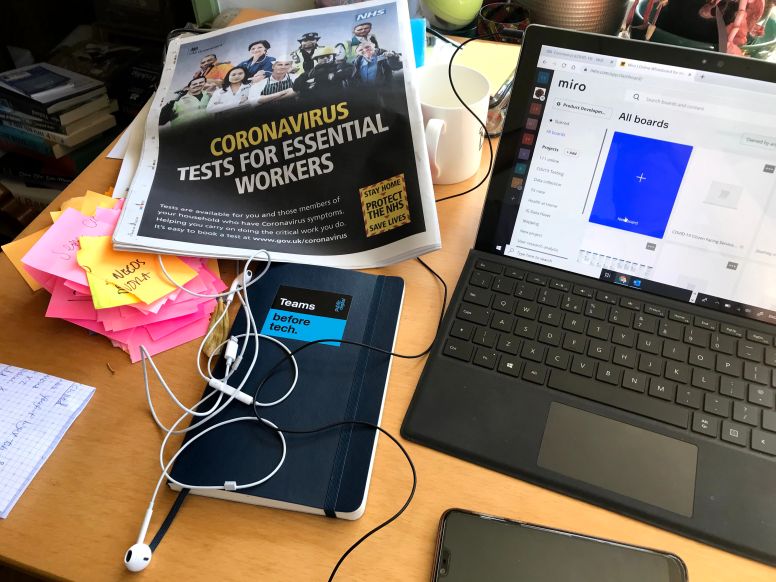 Desktop with newspaper showing "coronavirus tests for essential workers", notebook with sticker that says "teams before tech", torn up sticky notes, laptop with Miro open in browser