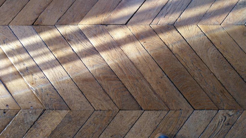 Strips of sunlight cast through a window cut across the herringbone pattern of an old wooden floor