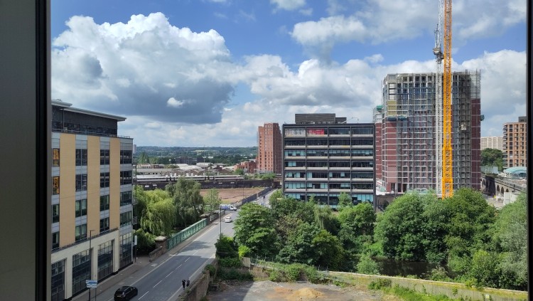 Looking down from a high window across a cityscape under a partially cloudy sky. A road runs over a tree-lined river, in the distance a train is travelling along a railway line between tall buildings and factories. Another tall building is under construction next to an old railway viaduct